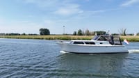 Een witte motorboot met een Nederlandse vlag vaart op een waterweg met groene oevers onder een blauwe lucht. — Valk Sport