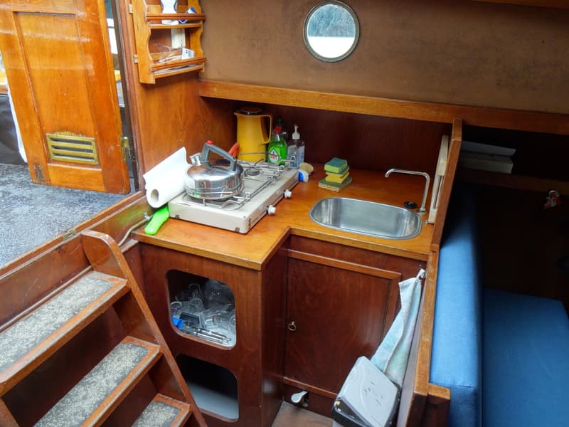 Een houten bootinterieur met een kombuis, trap, patrijspoort en blauwe kussens.