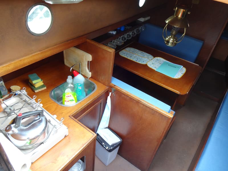 De foto toont een kajuit met een kombuis aan de linkerkant en een zithoek met tafel aan de rechterkant.