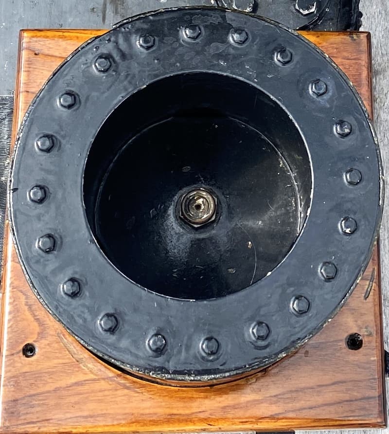 Een close-up van een zwart, rond metalen object met bouten, gemonteerd op een houten vierkante basis.