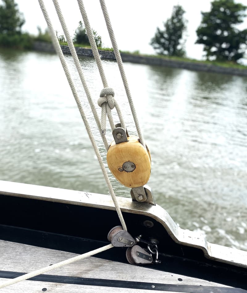 Een houten blok met touwen en twee kleinere blokken op een boot, met water en landschap op de achtergrond.