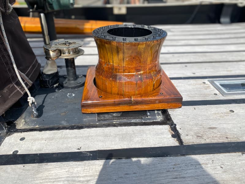 Een houten kaapstander met een metalen bovenrand en een lier op een houten dek.