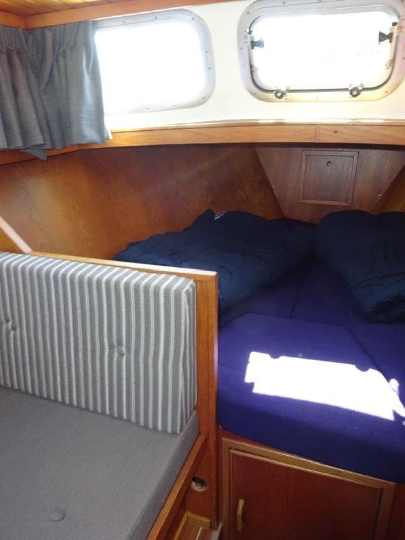 De foto toont een kajuit met houten wanden, twee patrijspoorten, een bank met gestreepte rugleuning en een bed met…