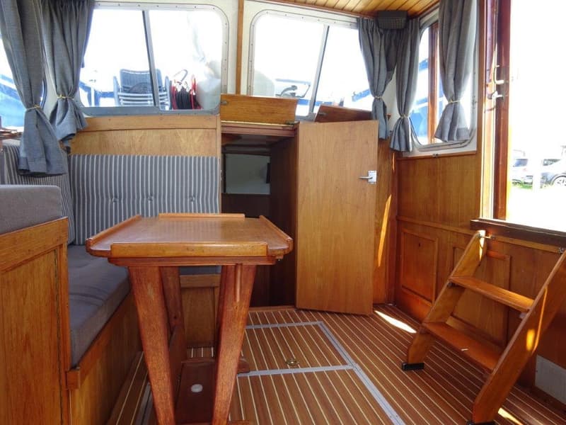 Een interieur van een boot met houten lambrisering, een tafel, banken met gestreepte kussens, ramen met gordijnen en een…