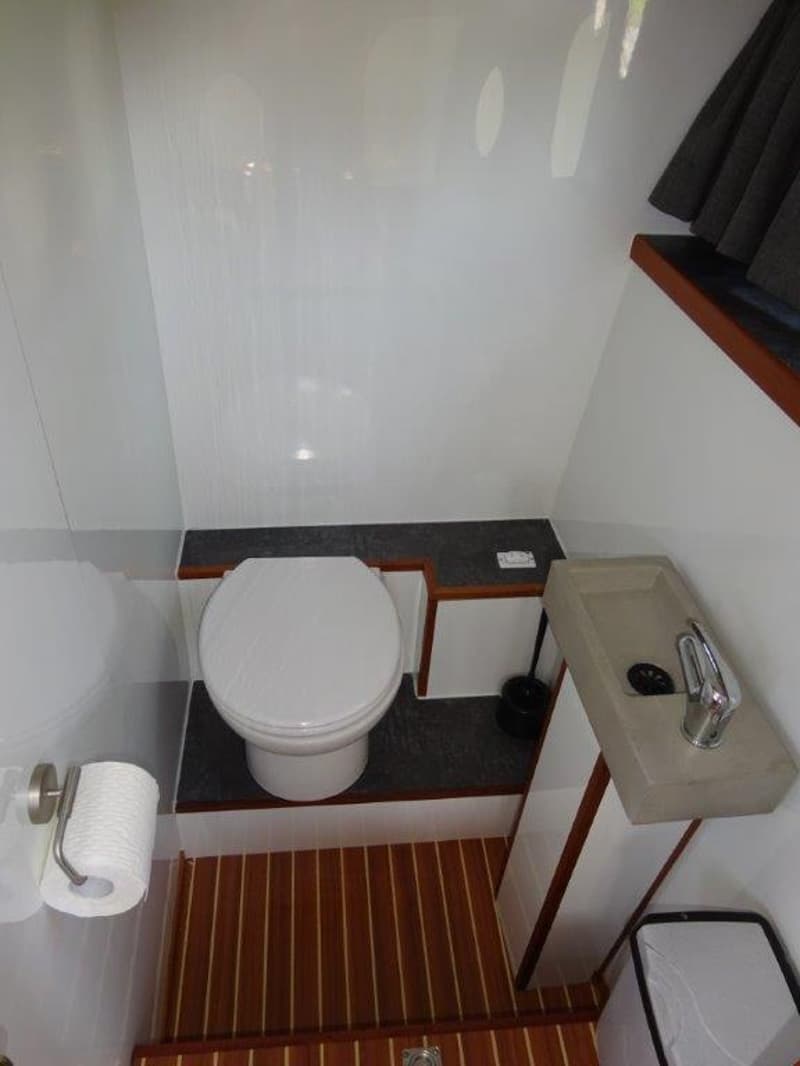 Een toiletruimte met een wit toilet, een grijze wasbak met kraan, een toiletrolhouder en een houten vloer.