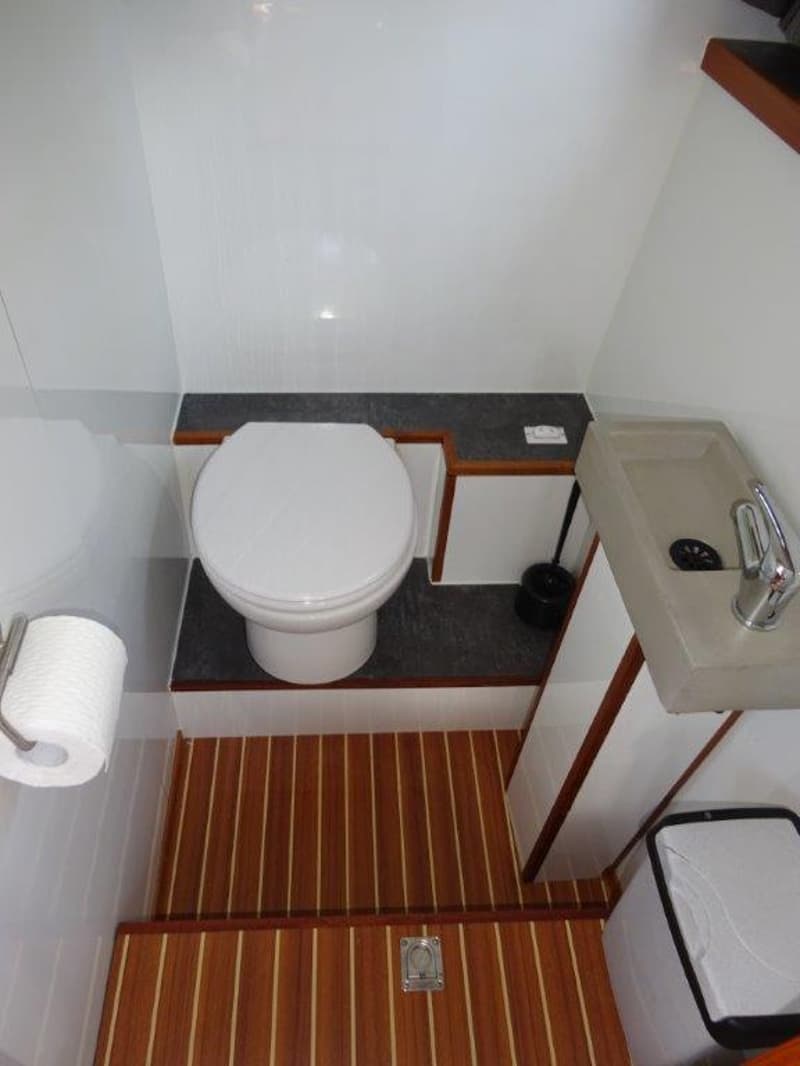 Een witte toiletruimte met een toilet, wastafel, toiletrolhouder en een houten vloer.