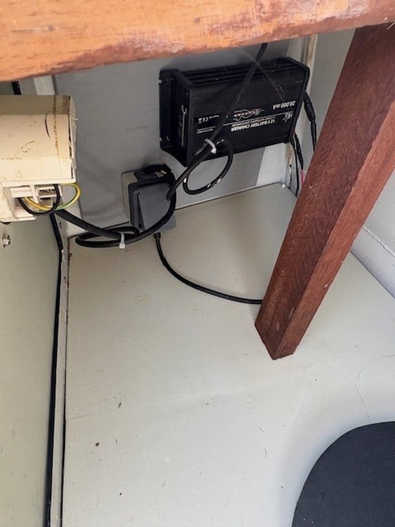 Een zwarte acculader met kabels is aan de muur bevestigd, met een witte doos en een houten balk ernaast.