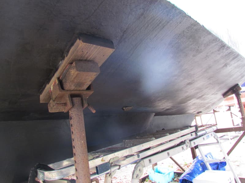 De onderkant van een donkergrijze boot op een metalen bok, met een ladder en blauw zeil op de grond. — Curtevenne 950 GSAK