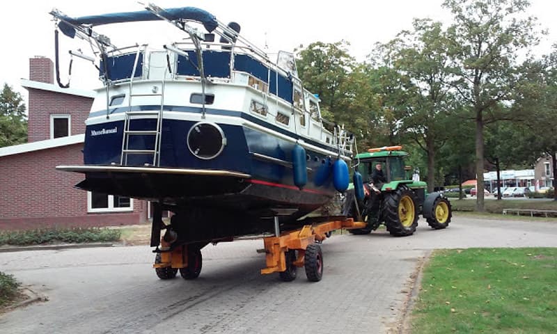 Een blauw-witte boot met de naam "Musselkanal" wordt op een trailer door een groene tractor getrokken. — Curtevenne 950 GSAK