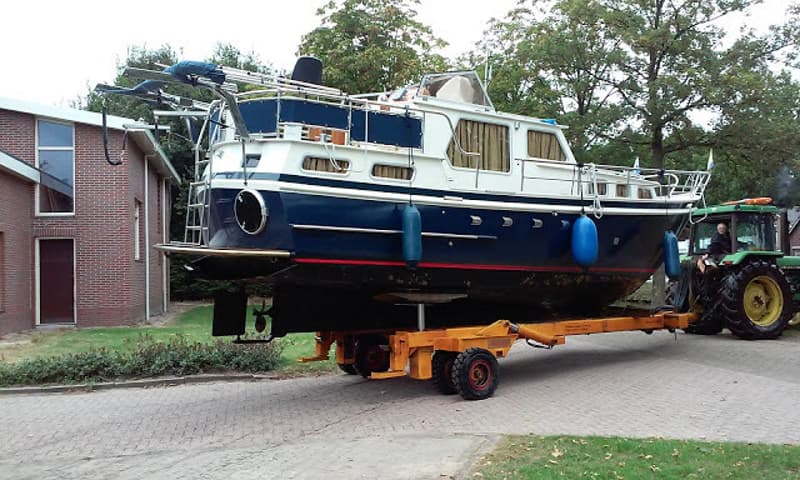 Een blauw-witte boot op een oranje trailer wordt getrokken door een groene tractor, met een man aan het stuur.