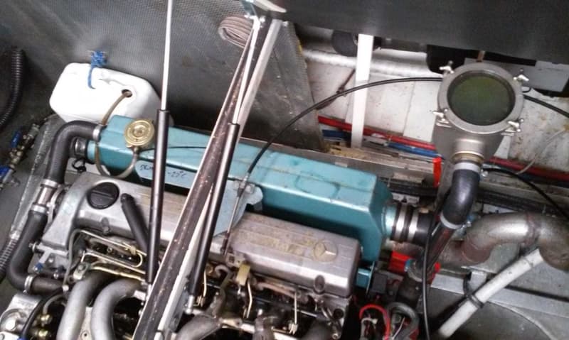 Een close-up van een motorruimte met een Mercedes-Benz motor, slangen, leidingen en een wit reservoir.
