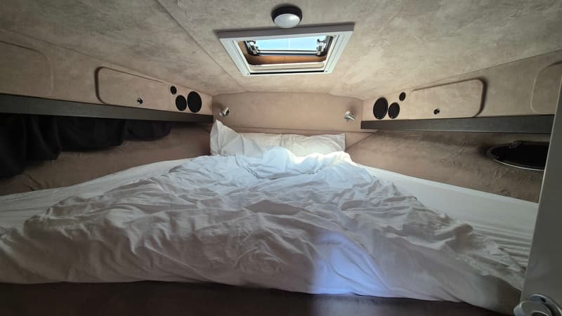 Een kajuit met een opgemaakt bed met wit beddengoed, een dakraam en ingebouwde luidsprekers en verlichting.
