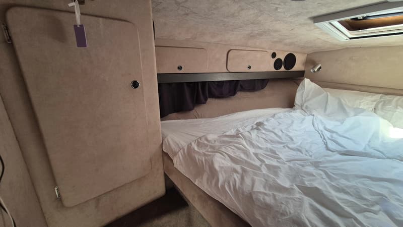 Een kajuit met een bed met wit beddengoed, opbergruimte, luidsprekers, een leeslampje en een dakraam.