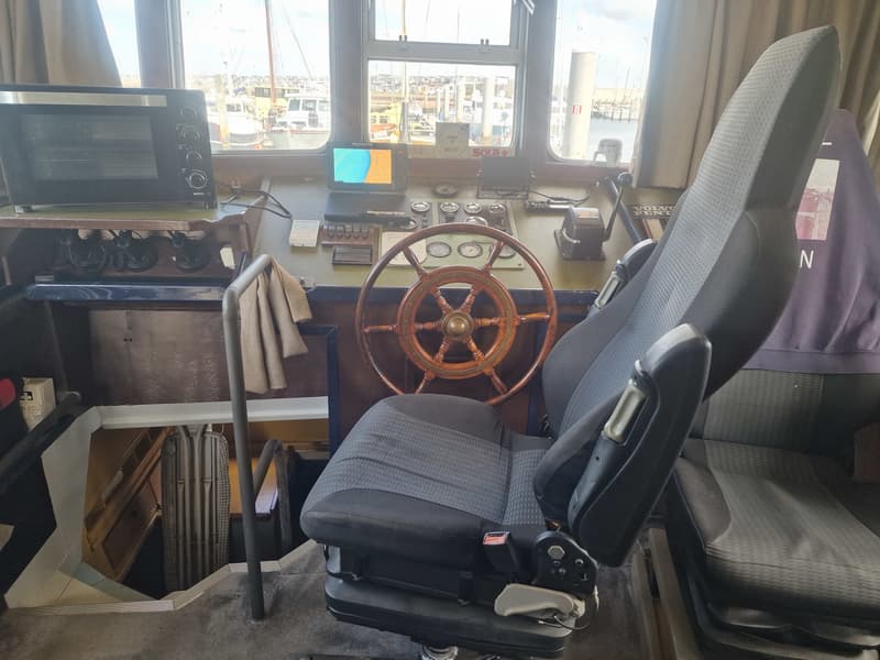 Een stuurhut met een houten stuurwiel, een Raymarine-scherm, een oven en een grijze stoel.