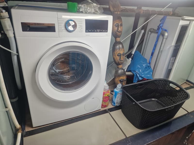Een witte Bosch wasmachine staat op een houten vloer, met wasmiddel en een wasmand ernaast.