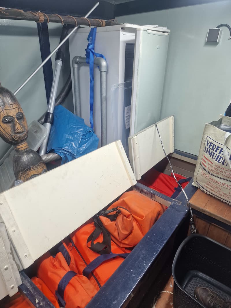 Een rommelige ruimte met een open koelkast, een houten beeld, oranje reddingsvesten en een tas met "VERFFABRIEK IJMUIDEN"…