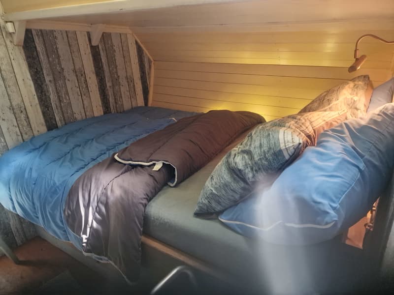 Een bed met blauwe en grijze dekens en kussens in een kajuit met houten wanden en een leeslampje.