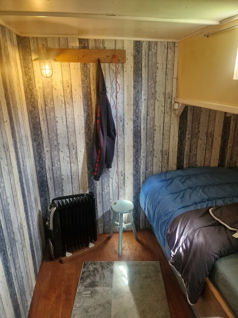 De kajuit heeft een bed met blauw en grijs beddengoed, een kachel, een krukje, een kapstok met een jas en een lamp.