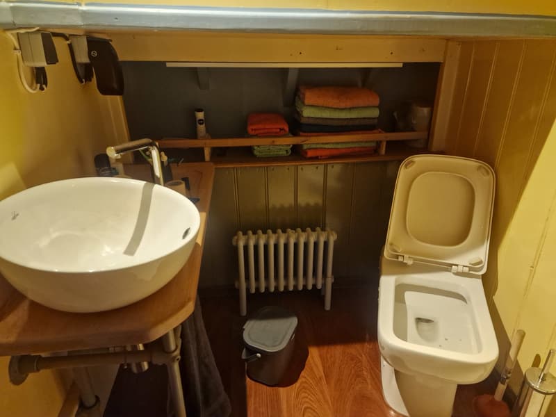 Een badkamer met een wit toilet, een witte wastafel op een houten blad, een radiator en handdoeken op planken.