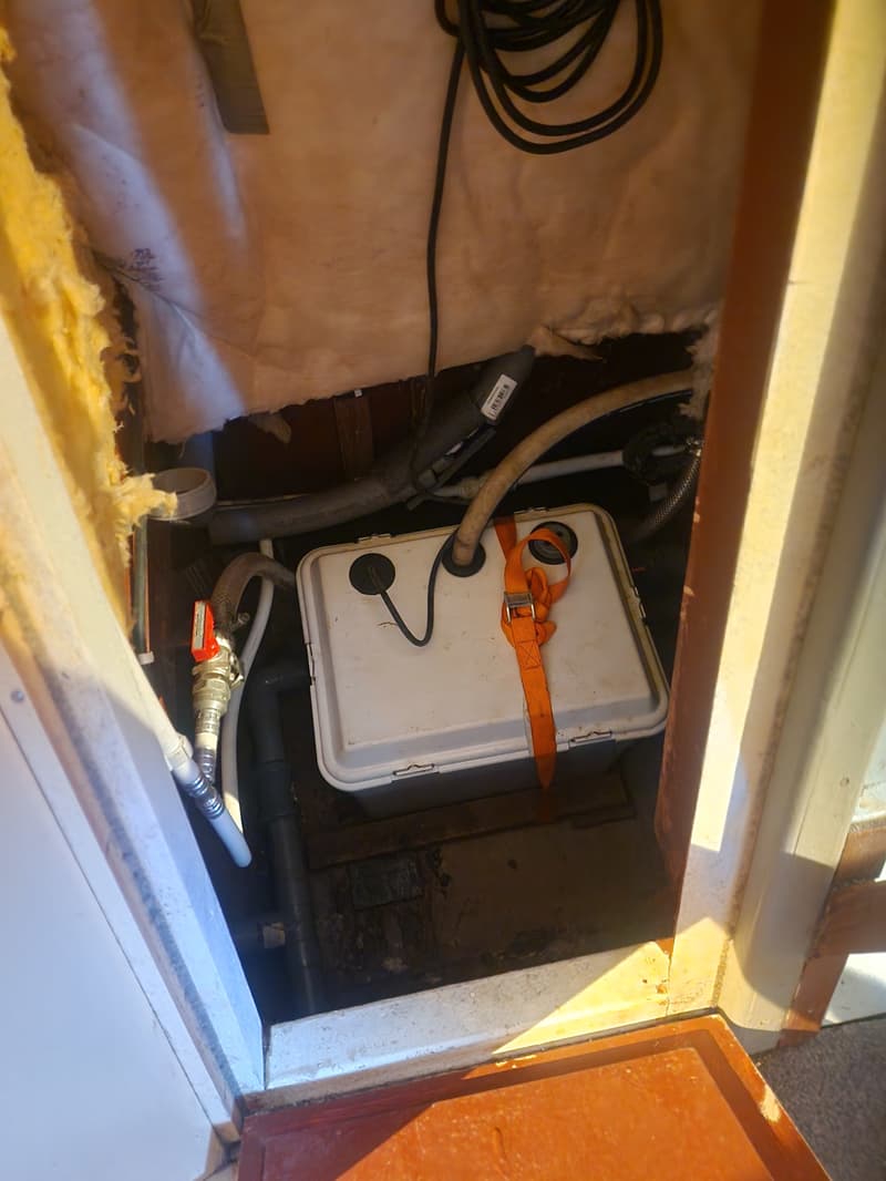 Een witte opbergbox met een oranje spanband, omringd door leidingen en isolatiemateriaal.
