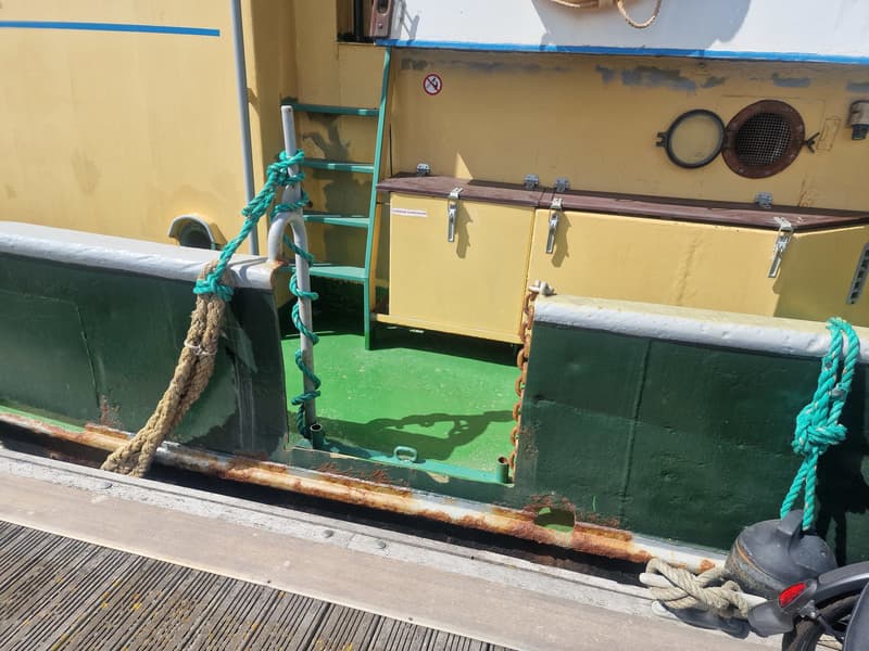 Een boot met een geel en groen dek, een ladder, touwen, een patrijspoort en een opslagkist met de tekst "VLOEIBAAR…