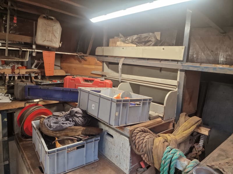 Een rommelige opslagruimte met gereedschap, touwen, jerrycans en kratten onder een TL-lamp.