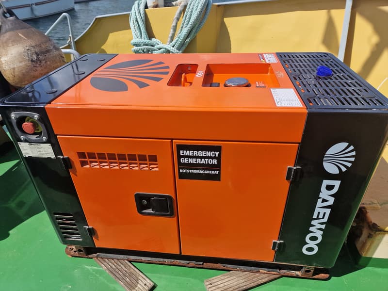 Een oranje en zwarte Daewoo noodstroomaggregaat staat op een groen dek, met een stootwil en touwen op de achtergrond.