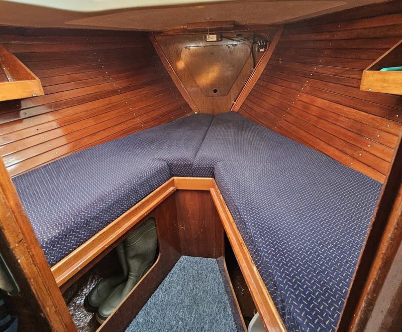 De foto toont het interieur van een bootkajuit met houten wanden en blauwe gestoffeerde kussens die een V-vormige…