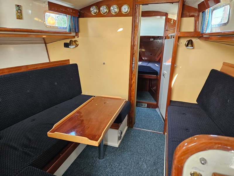 De foto toont het interieur van een boot met twee banken met donkerblauwe kussens, een houten tafel en een doorgang naar…