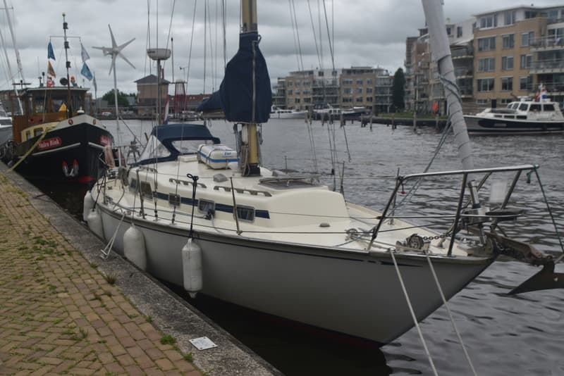 Een witte zeilboot met blauwe accenten ligt afgemeerd aan een kade, met een donkere boot ernaast. — Frans Maas Raider 35