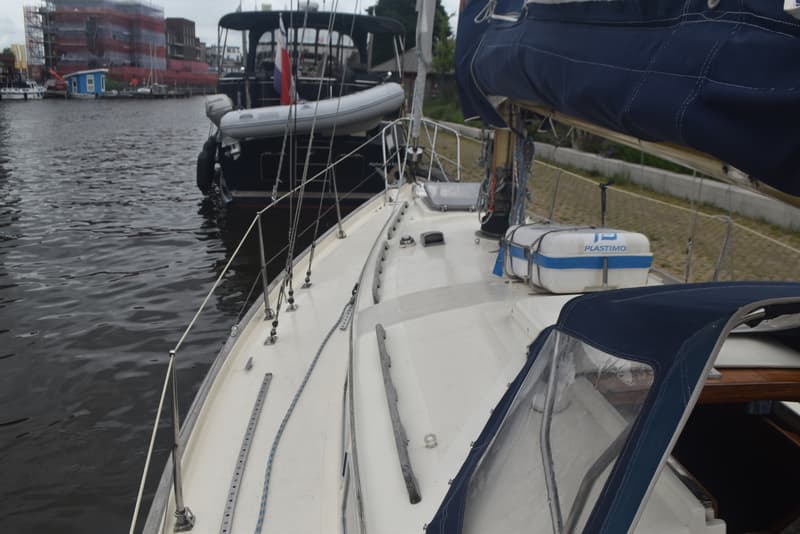Een zeilboot met een blauw zeil en een wit dek ligt in het water, met een andere boot en gebouwen op de achtergrond.