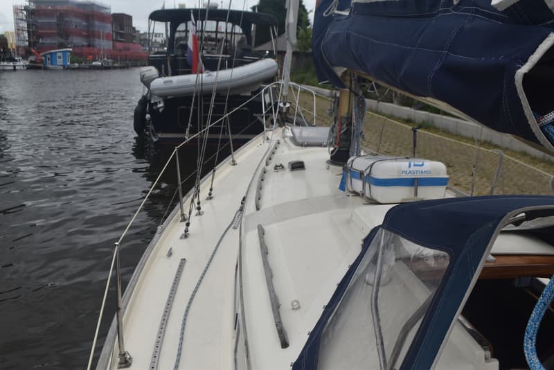 Een zeilboot met een blauw zeil en een reddingsvlot aan dek, met een andere boot en gebouwen op de achtergrond.