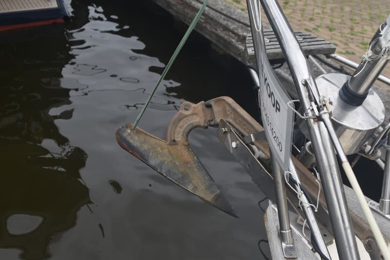 Een roestig anker hangt boven het water, met een wit bordje "TE KOOP" en een telefoonnummer eraan.