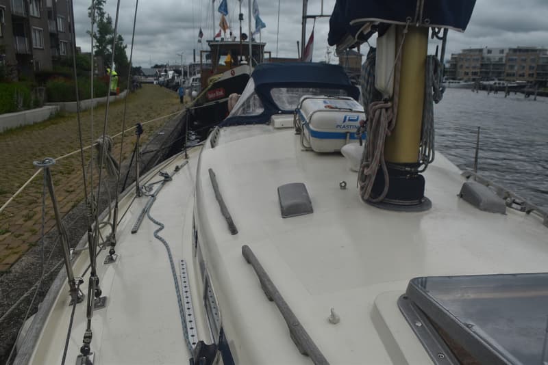 Een wit dek met een mast, touwen, een reddingsvlot, en een blauwe buiskap, met een kade en gebouwen op de achtergrond.