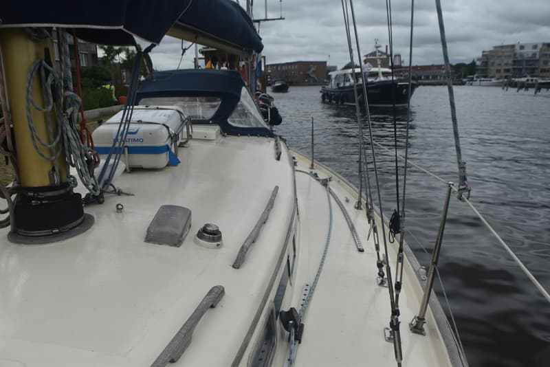 Een witte zeilboot met een blauwe buiskap en een reddingsvlot, liggend aan een kade met een andere boot op het water.