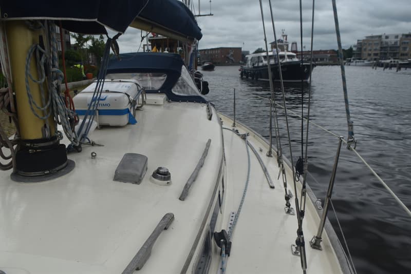 Een wit zeiljacht met een blauwe buiskap en een reddingsvlot aan dek, liggend in een haven.