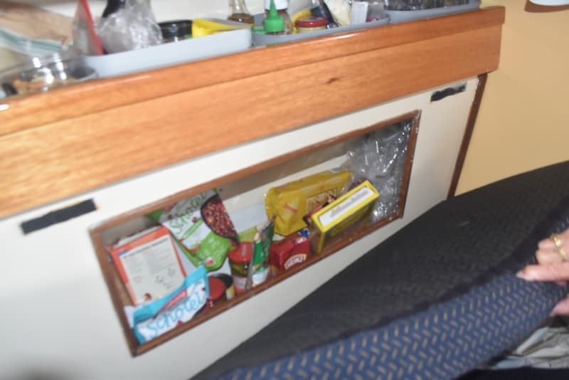 Een interieur van een boot met een houten rand, een open opbergvak met etenswaren en een donkerblauw kussen.