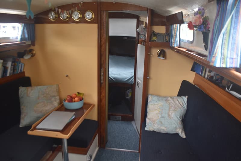 De foto toont het interieur van een bootkajuit met twee banken, een tafel, een doorgang naar een slaapruimte en diverse…