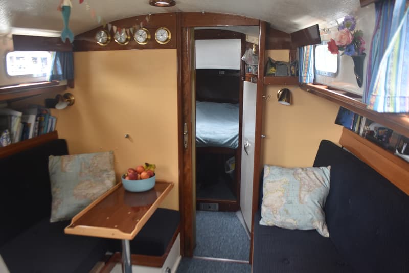 De foto toont een interieur van een boot met twee banken, een tafel met fruit, en een doorgang naar een slaapcabine.