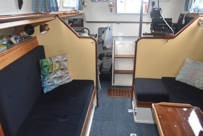 De foto toont het interieur van een boot met twee banken met donkerblauwe kussens, een tafel en een kombuis.