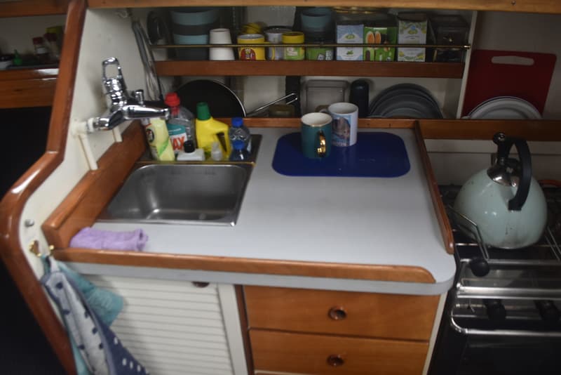 Een bootkeuken met een gootsteen, kraan, fornuis, waterkoker, theedoeken, en diverse keukengerei en voorraad.