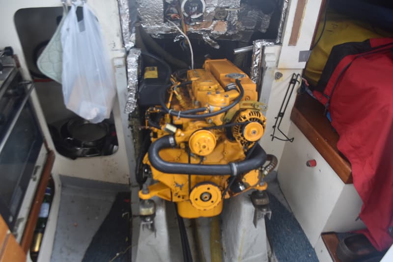 Een gele motor met zwarte slangen en riemen is zichtbaar in een bootinterieur met folie-isolatie en een rode jas.