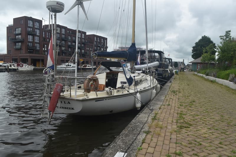 Een witte zeilboot met de naam "ILA S'ERBEEK" ligt aangemeerd aan een kade, met gebouwen op de achtergrond.