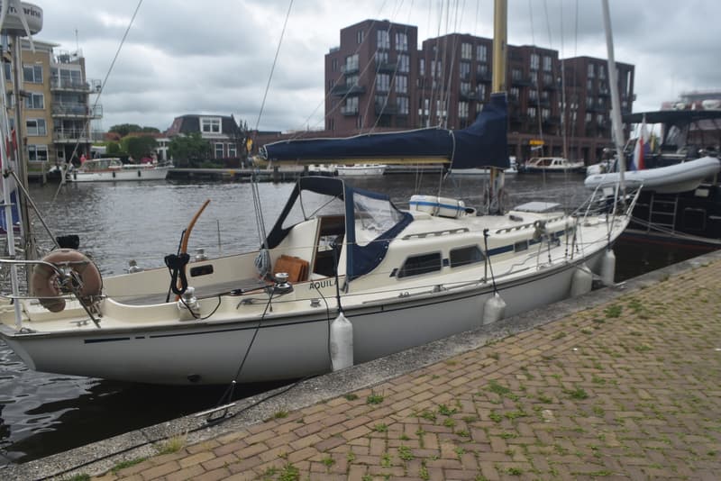 Een zeilboot met de naam "AQUILA" ligt aangemeerd aan een kade van bakstenen, met gebouwen op de achtergrond.