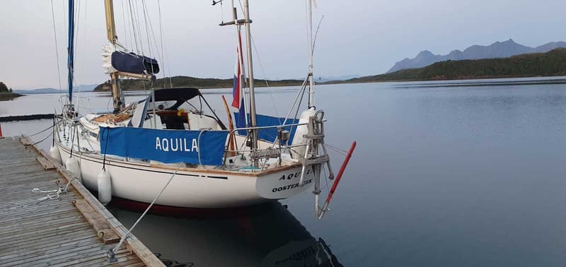 Een witte zeilboot met de naam "AQUILA" en "OOSTERBEEK" op de spiegel ligt afgemeerd aan een houten steiger.