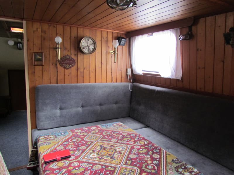 Een interieur van een boot met houten wanden en plafond, een grijze hoekbank, een tafel met een rood kleed en een raam met…