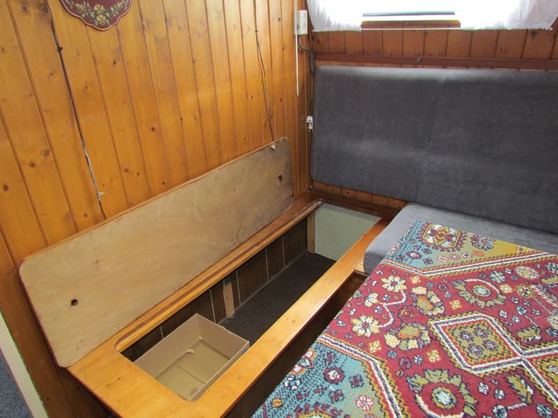 Een houten interieur met een open opbergruimte onder een bank met een grijs kussen en een kleurrijk kleed.