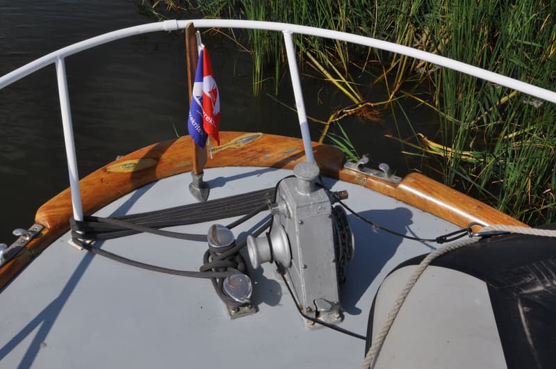 De boeg van een boot met een ankerlier, bolders, een vlag en een stootwil, met riet langs de waterkant.