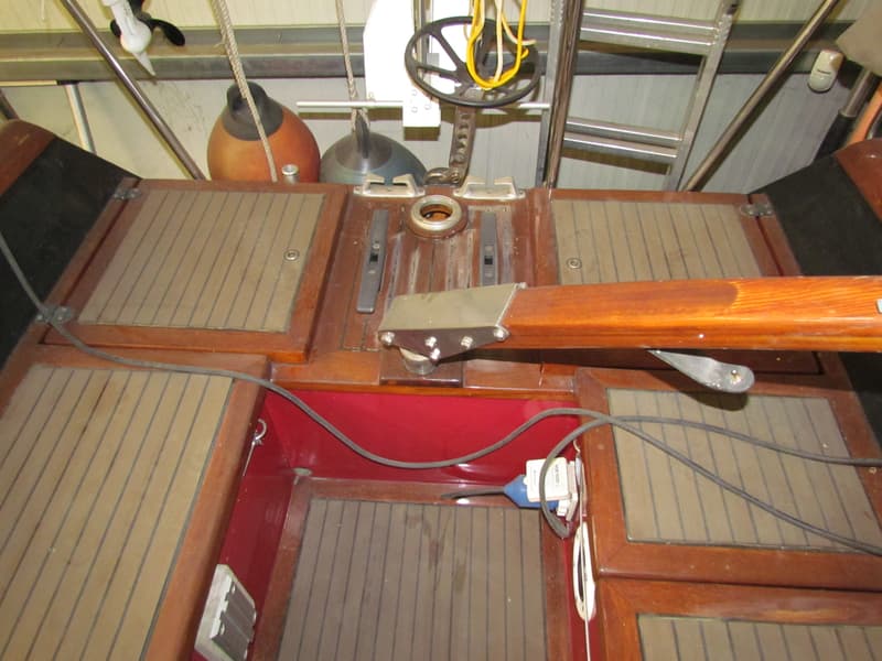 Een bovenaanzicht van een houten bootdek met een roer, fenders, touwen en een stuurwiel.
