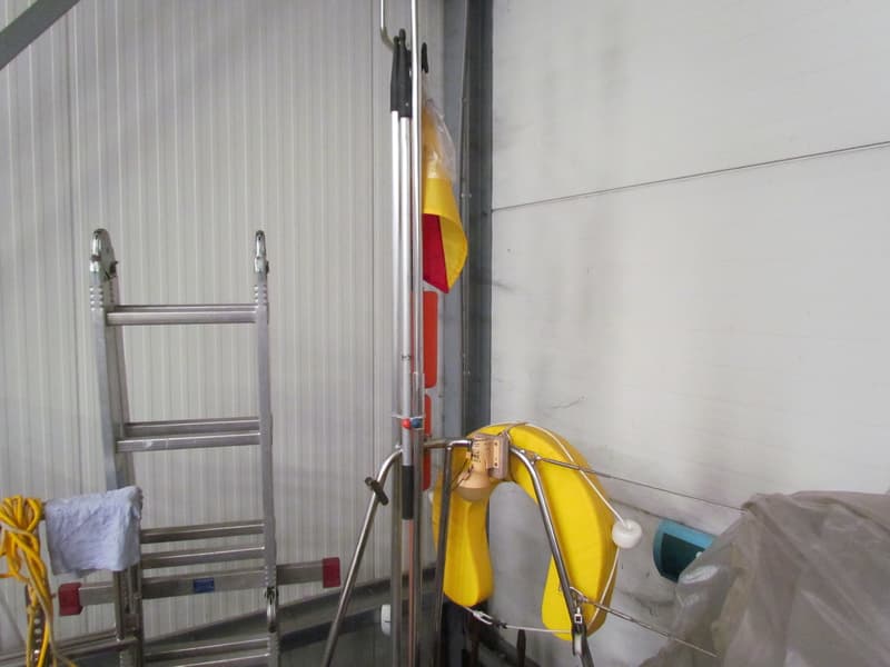 Een gele reddingsboei hangt aan een metalen rek, met een ladder en een vlag op de achtergrond.